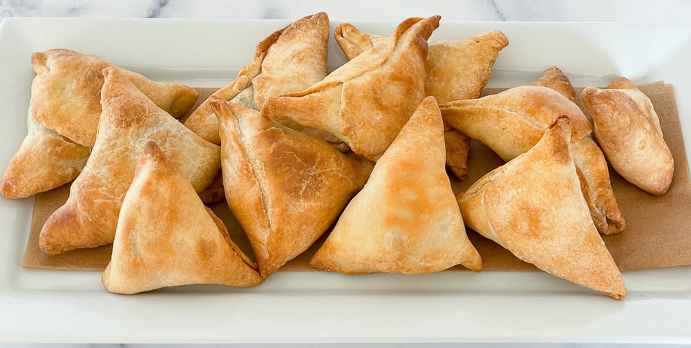 A rectangular white plate holds several golden-brown, triangular pastries arranged in a single layer on a brown paper strip.