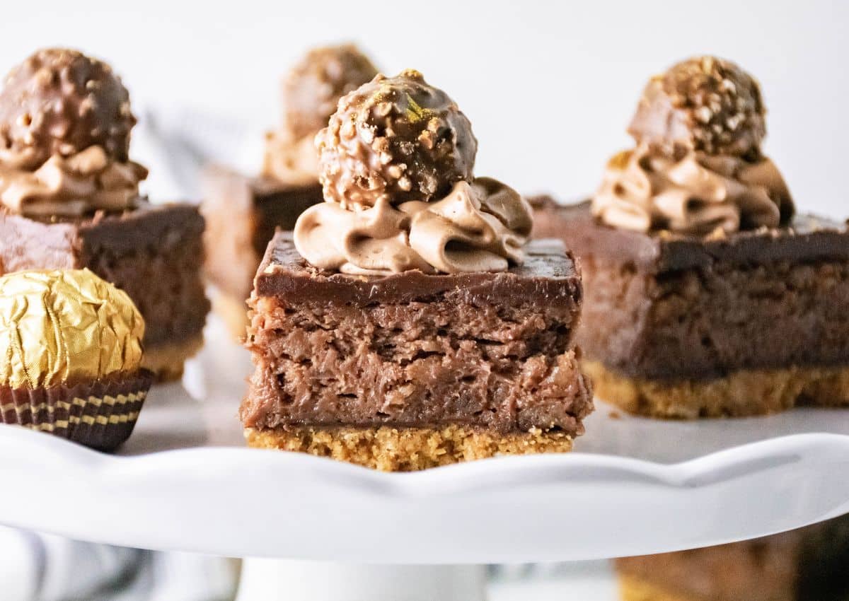 A close-up of chocolate dessert bars topped with chocolate frosting and a chocolate hazelnut candy, displayed on a white cake stand.