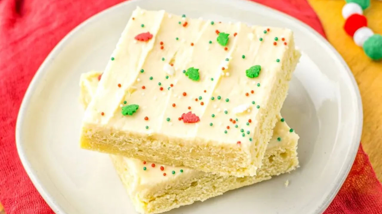 Two frosted sugar cookie bars with colorful sprinkles are stacked on a white plate, sitting on a red napkin.