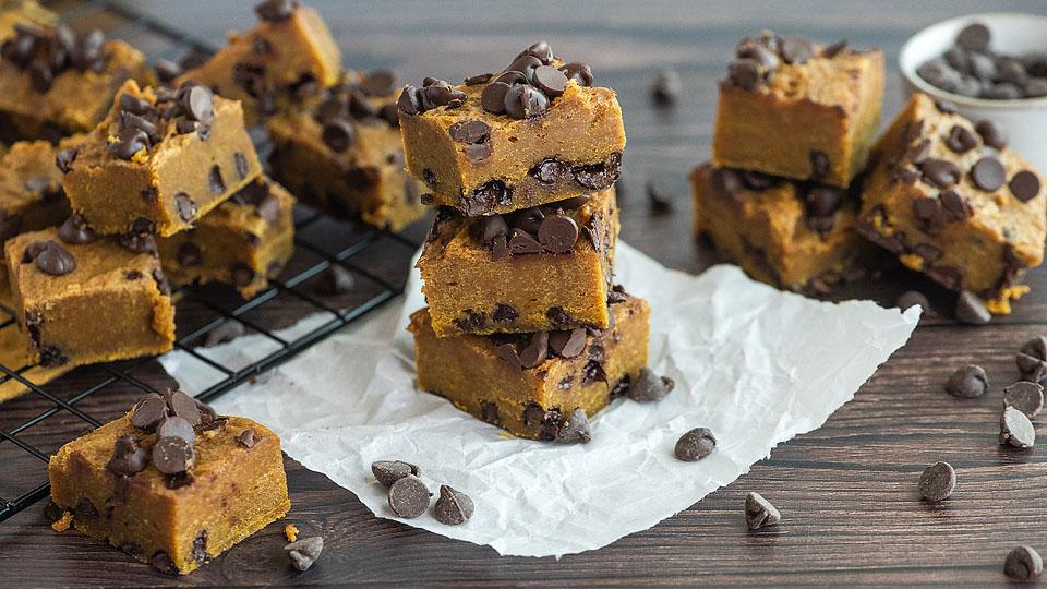 Squares of chocolate chip cookie dough fudge are stacked on parchment and a cooling rack, with scattered chocolate chips on a wooden surface.