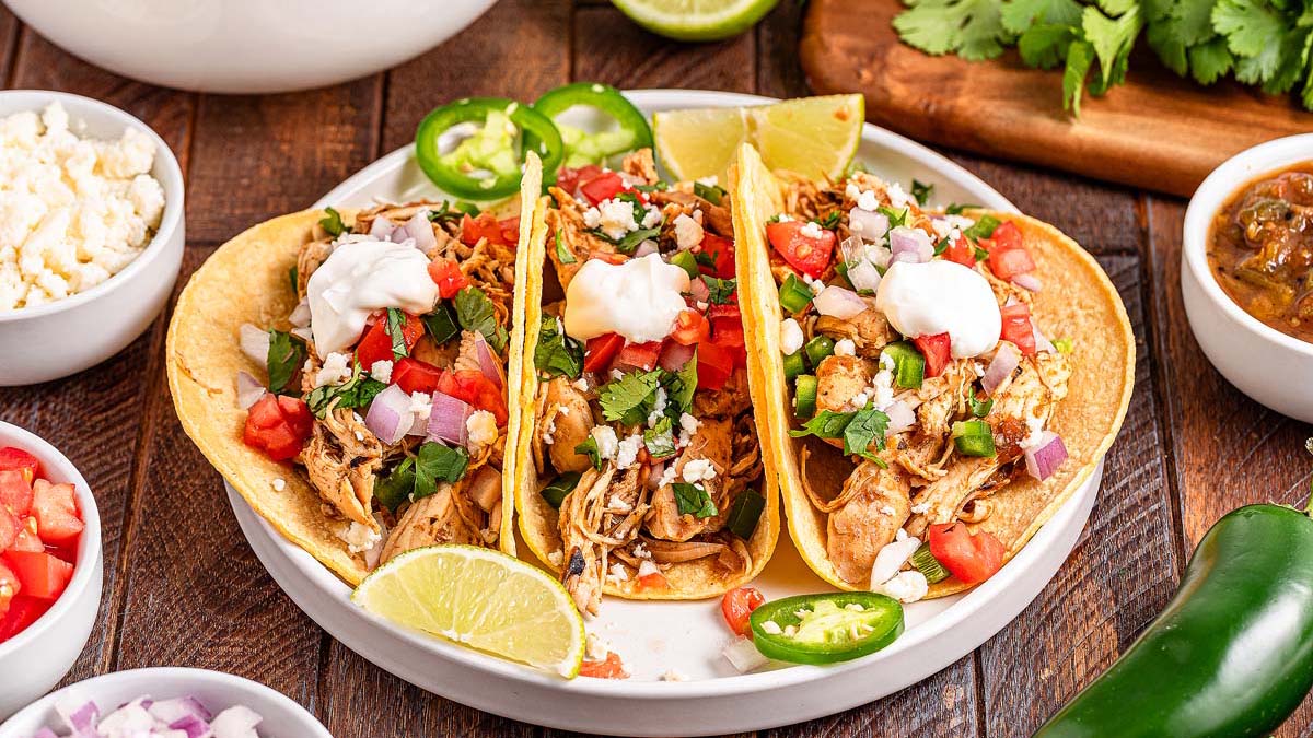 Three shredded chicken tacos in corn tortillas topped with diced tomatoes, onions, cilantro, jalapeรฑos, and sour cream, served with lime wedges on a white plate.