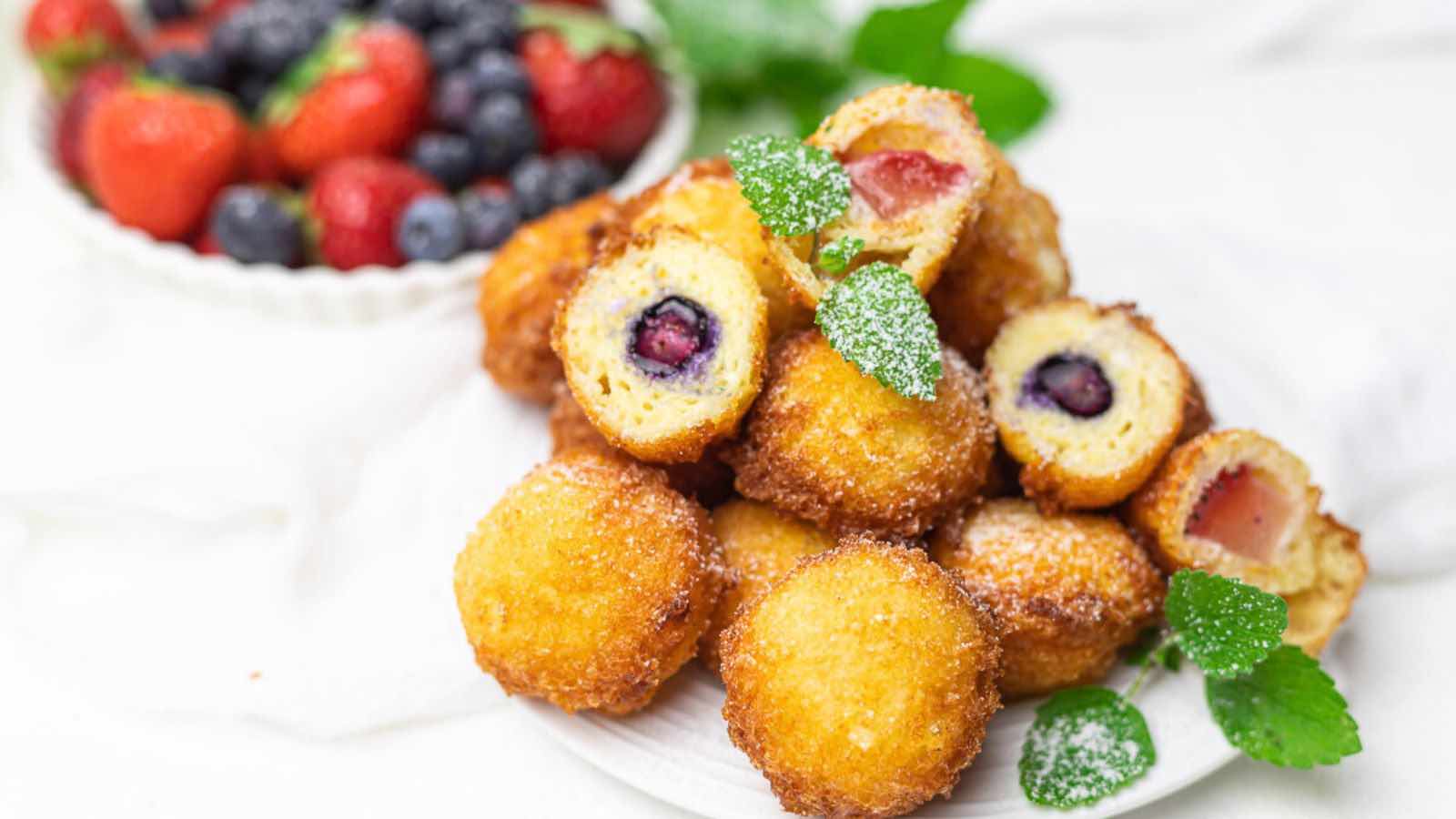 A plate of golden-brown fried dough balls with fruit filling, garnished with mint leaves and powdered sugar, with a bowl of fresh berries in the background.
