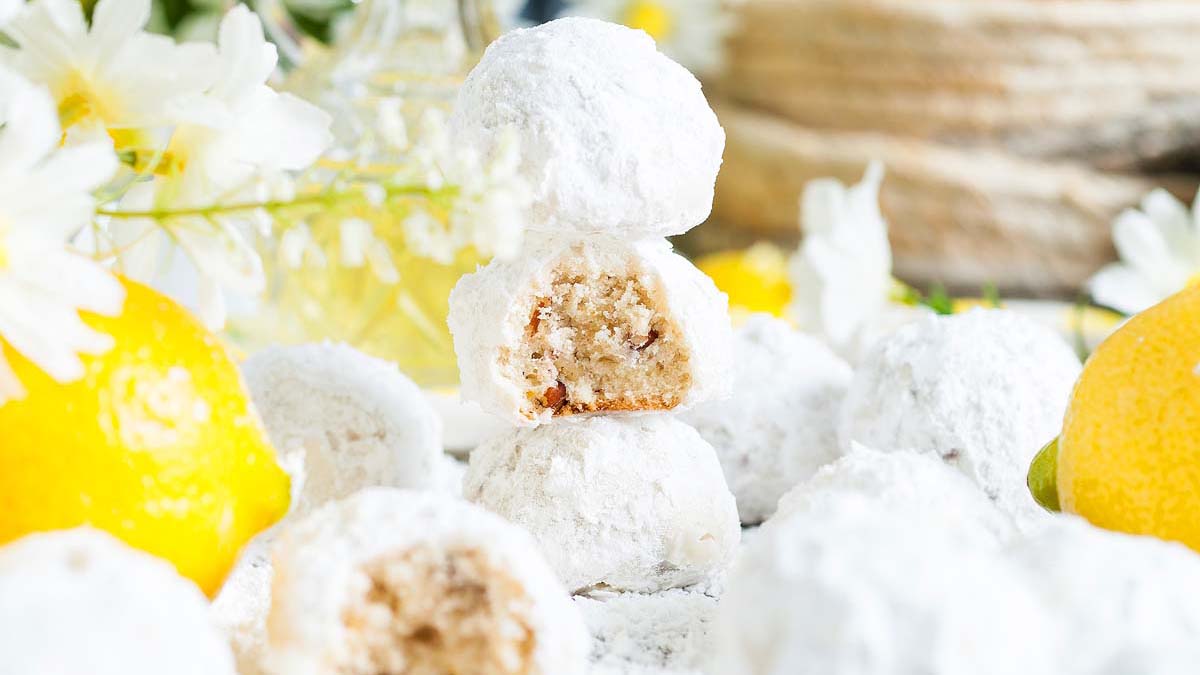 Powdered sugar-coated cookies are stacked with a bite taken out of the top one, surrounded by whole lemons and white flowers.