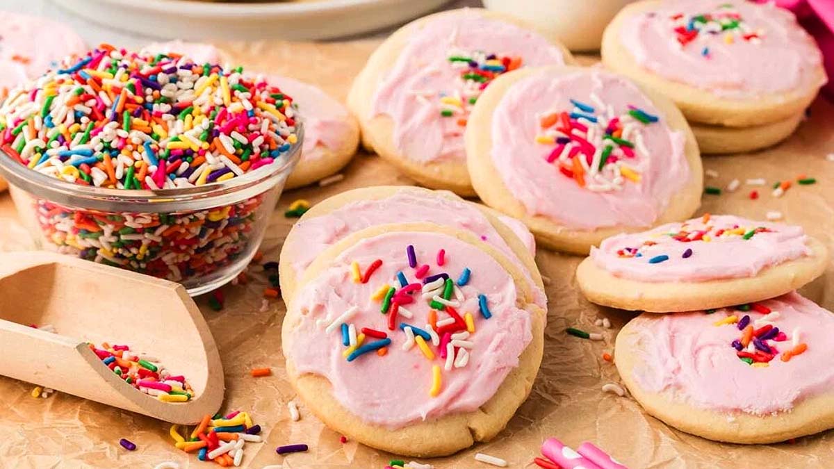 Sugar cookies with pink frosting and colorful sprinkles are arranged on parchment paper, with a small bowl and scoop of sprinkles nearby.