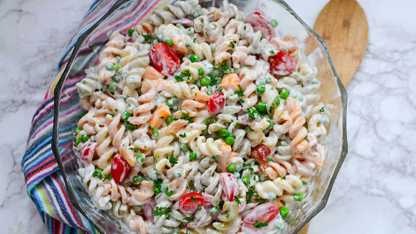 A glass bowl of creamy pasta salad with rotini noodles, cherry tomatoes, green peas, and herbs, next to a wooden spoon on a striped cloth.