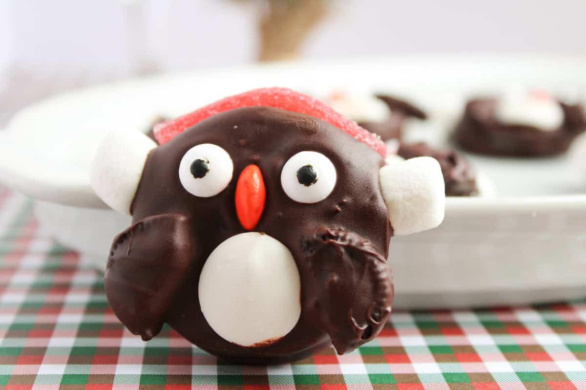 A chocolate-covered cookie decorated to look like a penguin with white and orange candy details, sitting on a plaid tablecloth near a white plate.