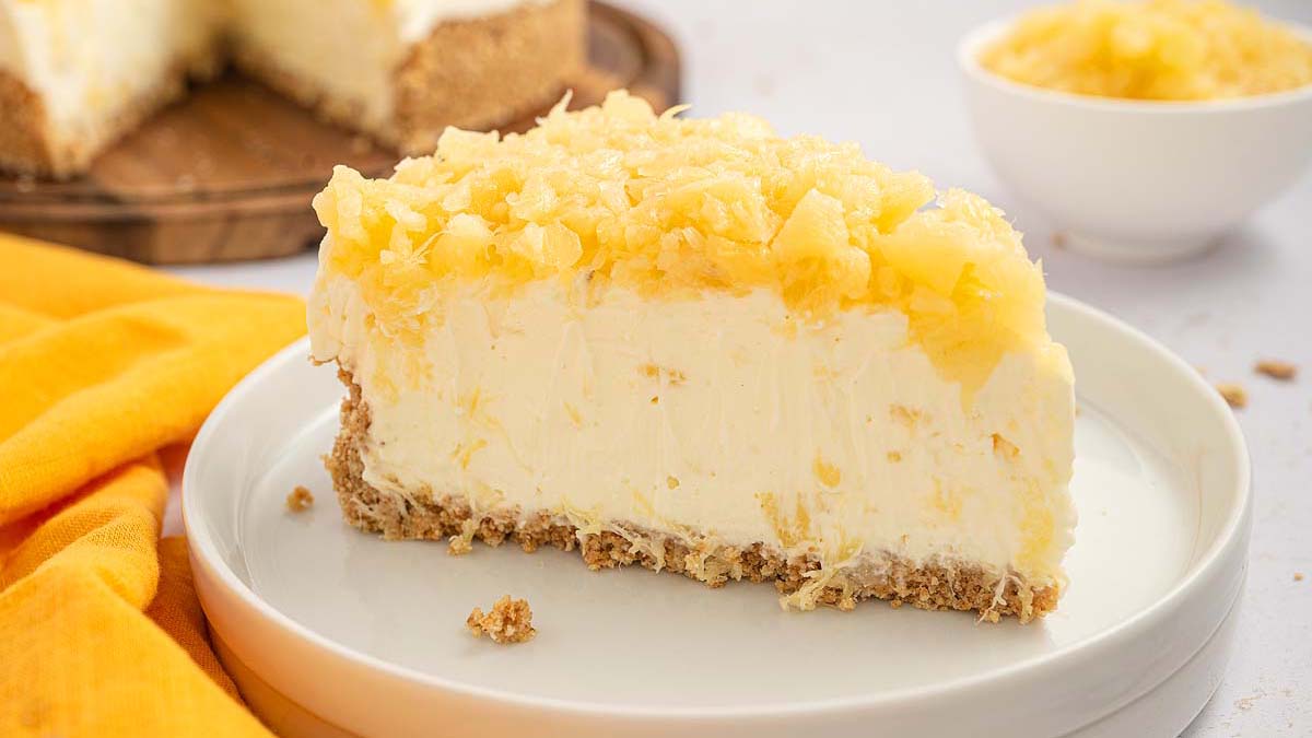 A slice of pineapple-topped cheesecake with a crumbly crust on a white plate, with a bowl of pineapple and more cheesecake in the background.