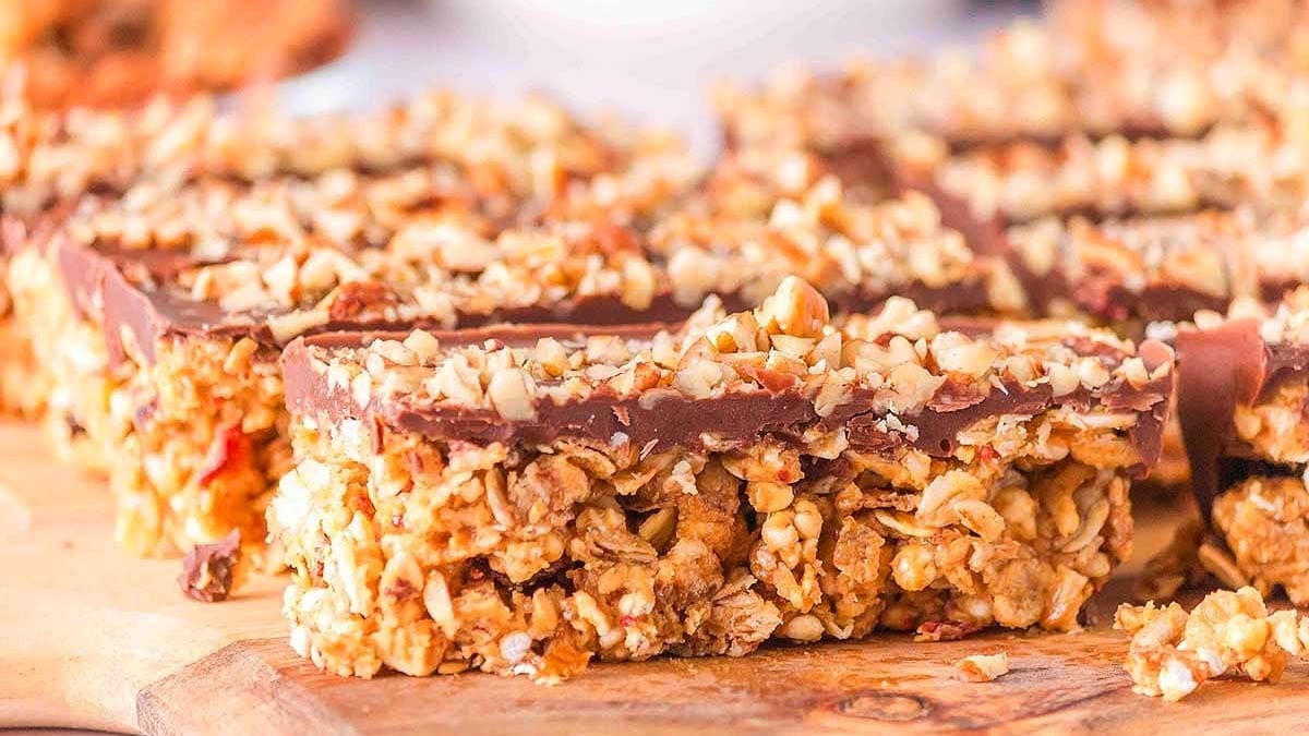 A close-up of homemade granola bars topped with chopped nuts and a layer of chocolate, arranged on a wooden surface.