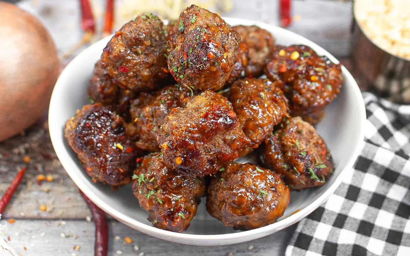 A white bowl filled with glazed meatballs, garnished with herbs, sits on a table next to a checkered napkin and assorted spices.