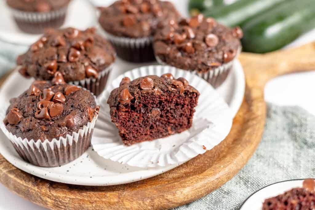 A plate of chocolate chip muffins, one with a bite taken out, sits on a wooden tray with a green cloth and zucchinis in the background.