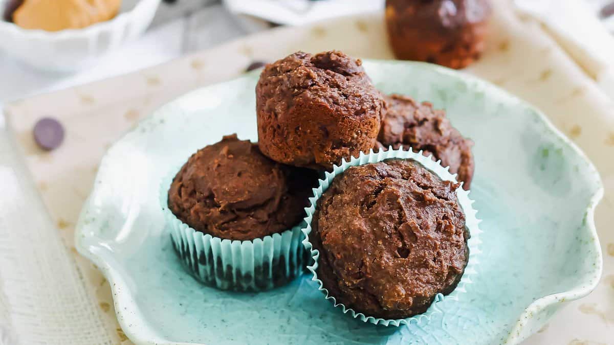 Four chocolate muffins in blue paper liners are arranged on a light blue ceramic plate. A blurred muffin and bowl are visible in the background.