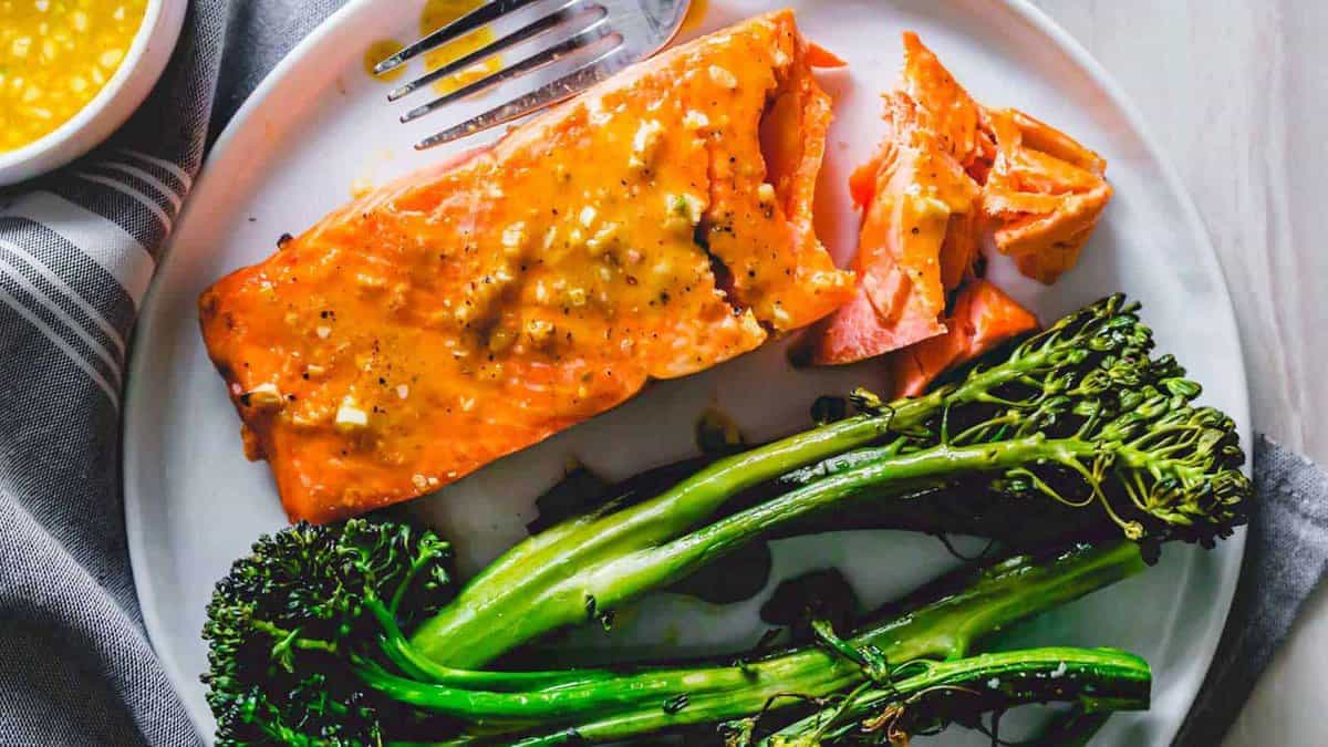 A plate with a cooked salmon fillet topped with sauce and a serving of broccolini, with a fork on the side.