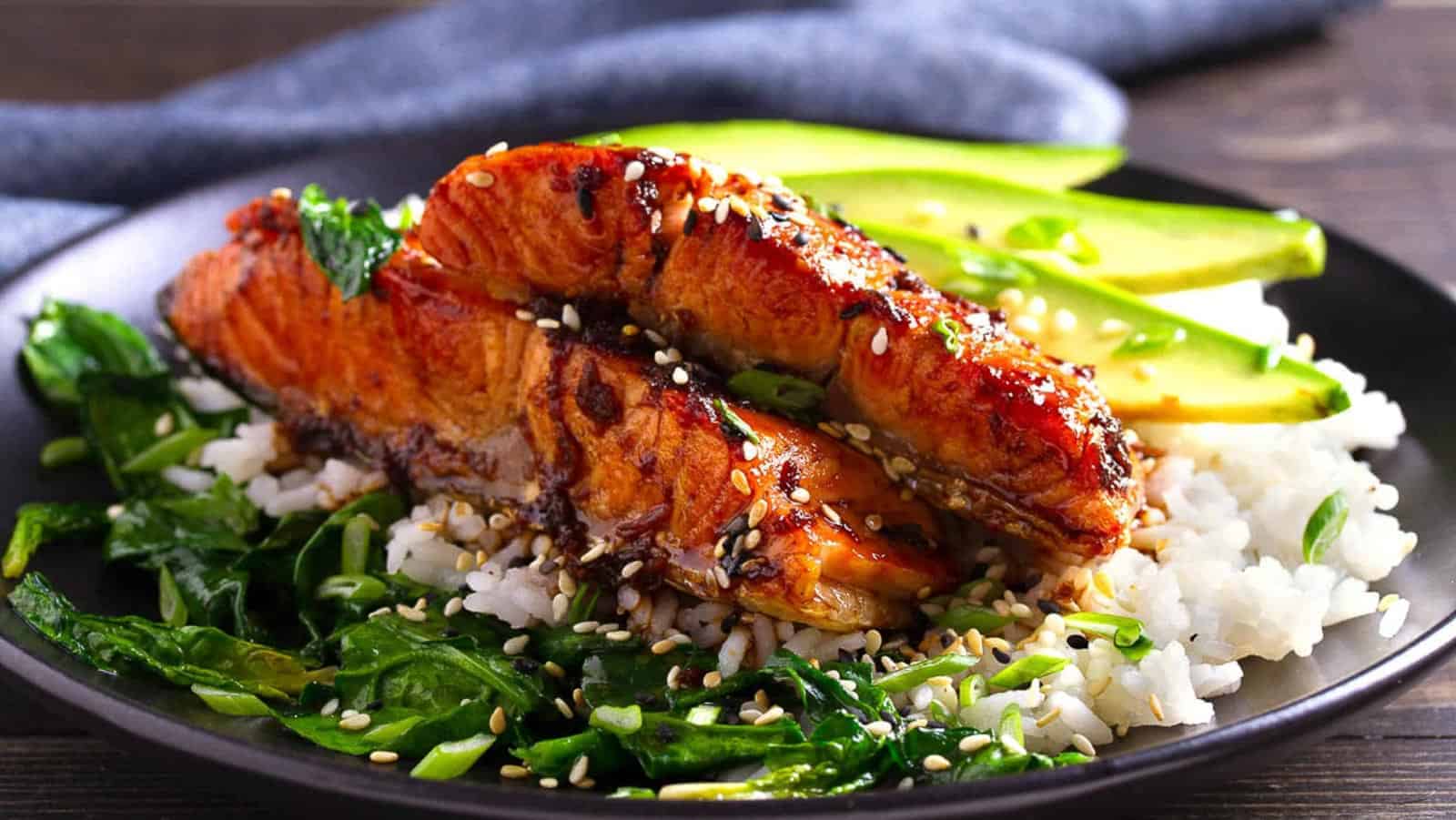 Grilled salmon fillets served on white rice with sautรฉed spinach, sliced avocado, and sprinkled sesame seeds on a black plate.