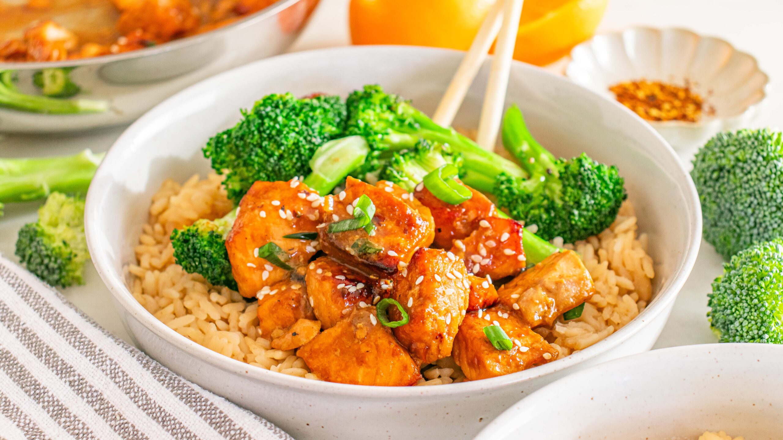 A bowl of brown rice topped with pieces of glazed chicken, broccoli florets, chopped green onions, and sesame seeds, with chopsticks resting on the bowl.