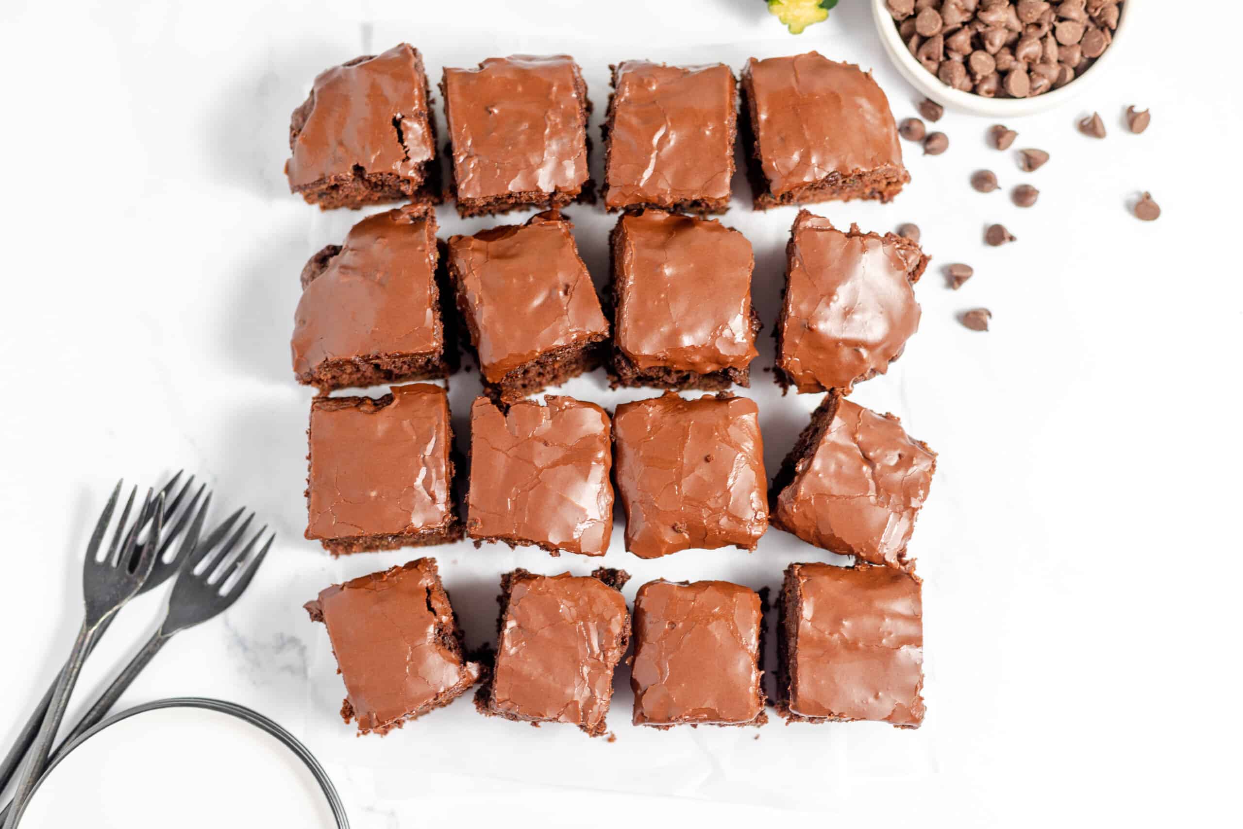 Fifteen chocolate brownies with glossy frosting arranged in a grid on parchment paper, with a bowl of chocolate chips and three forks nearby.