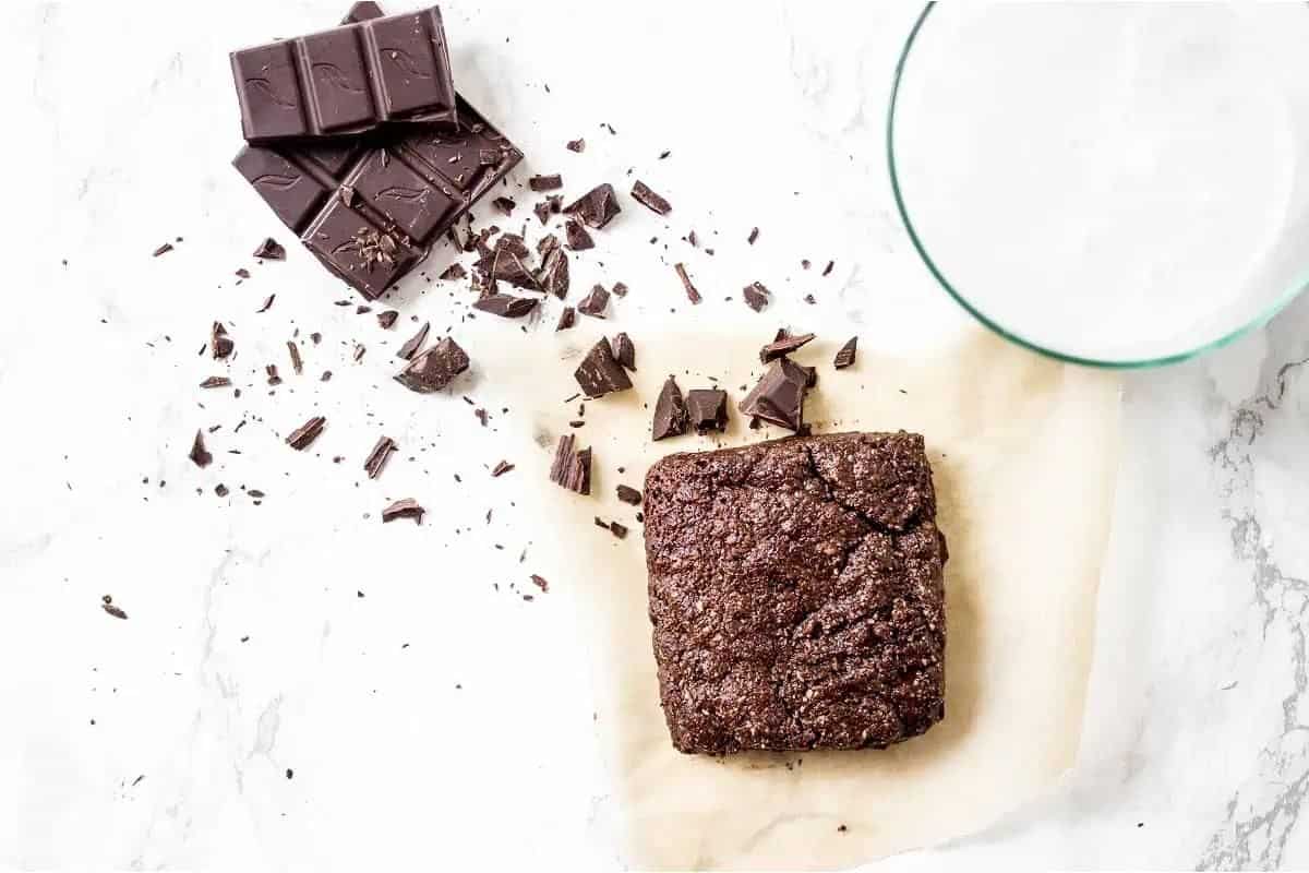 A chocolate brownie on parchment paper next to a pile of chocolate chunks and a glass of milk on a white marble surface.