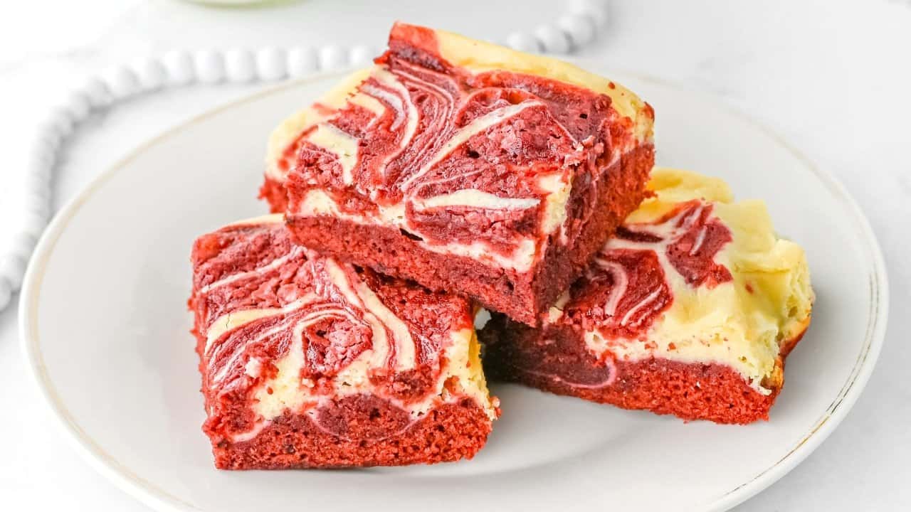 Three pieces of red velvet cheesecake brownies with a marbled cream cheese swirl are stacked on a white plate.