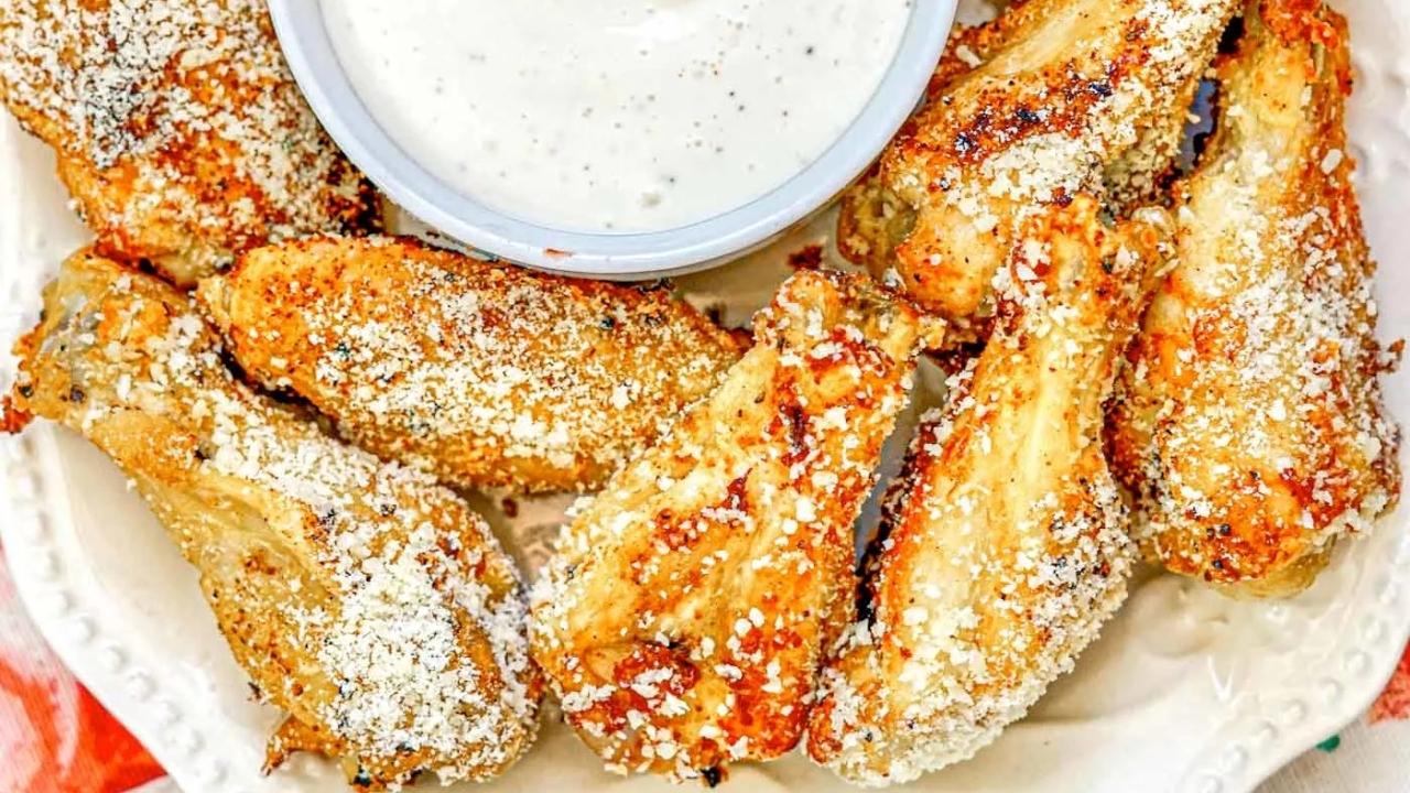 A plate of seasoned chicken wings topped with grated cheese, arranged around a bowl of white dipping sauce.