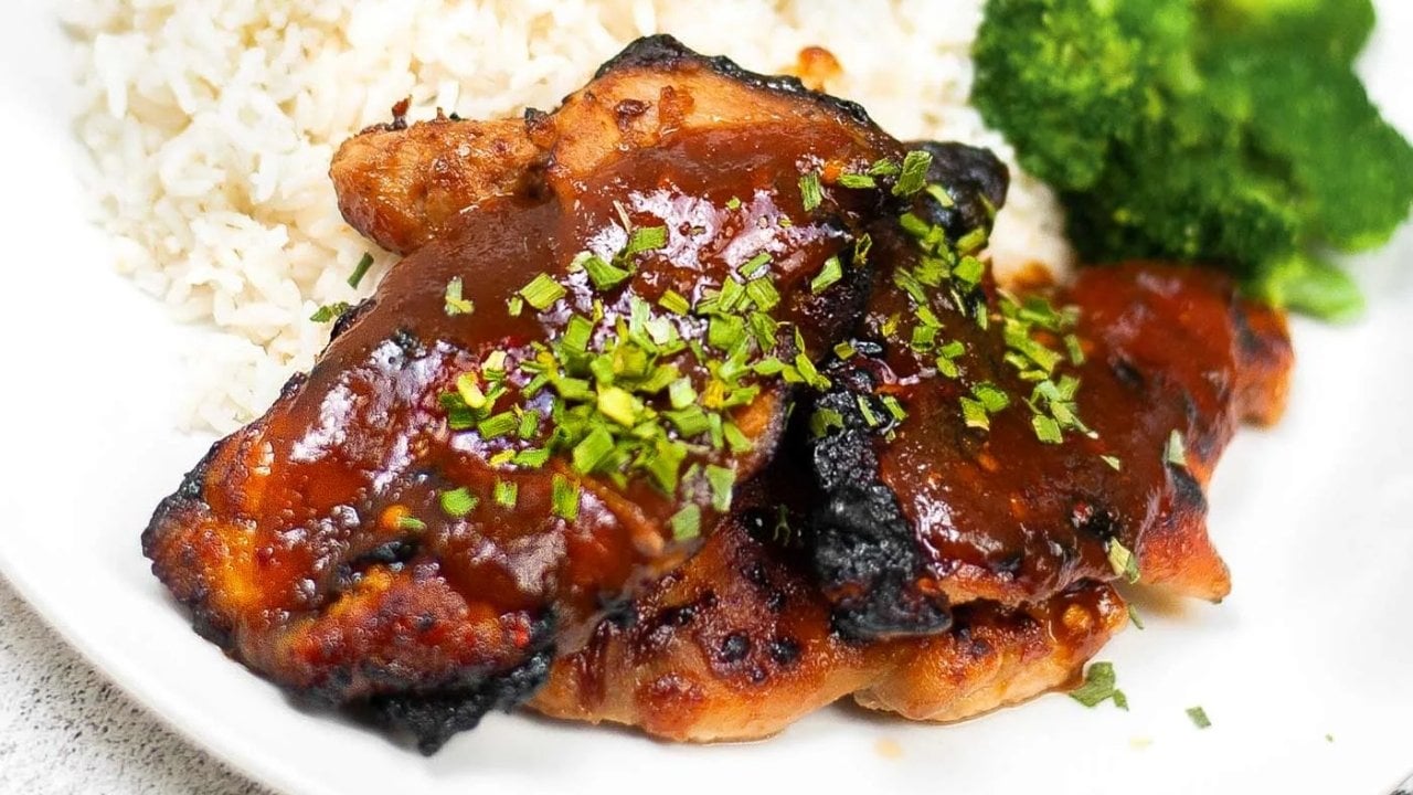 Two pieces of glazed grilled chicken garnished with chopped herbs, served with white rice and broccoli on a white plate.