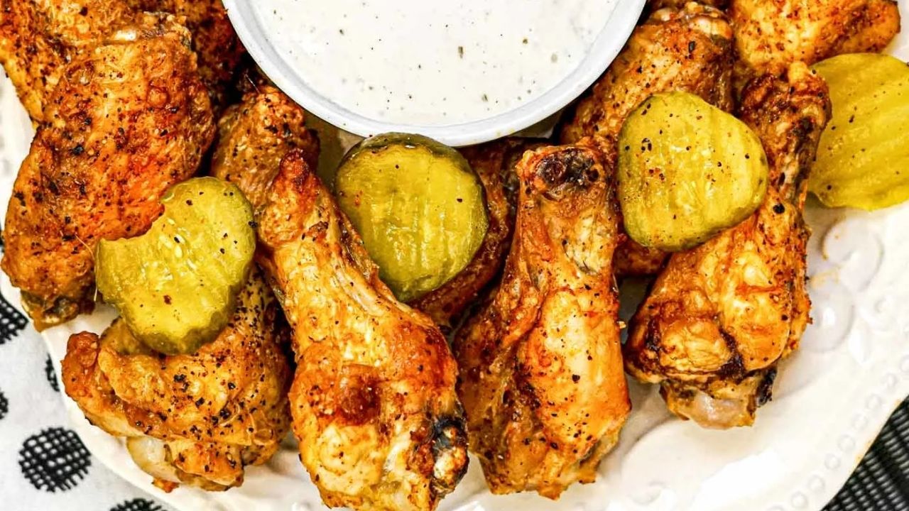 A plate of seasoned chicken wings garnished with pickle slices, served with a cup of creamy dipping sauce.