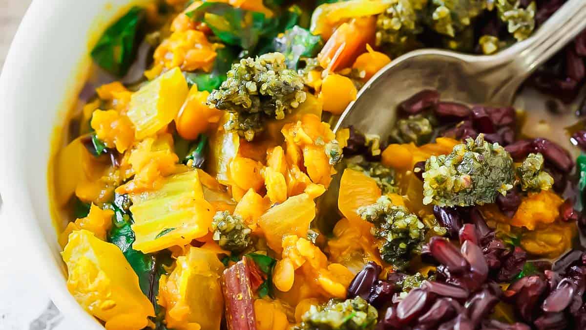 A close-up of a vegetable stew with yellow squash, leafy greens, broccoli, and purple rice in a white bowl with a spoon.