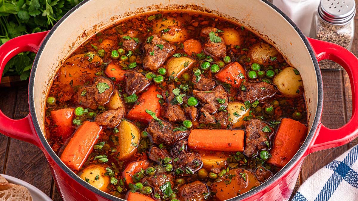 A red pot filled with beef stew containing chunks of beef, carrots, potatoes, peas, and herbs in a brown broth.