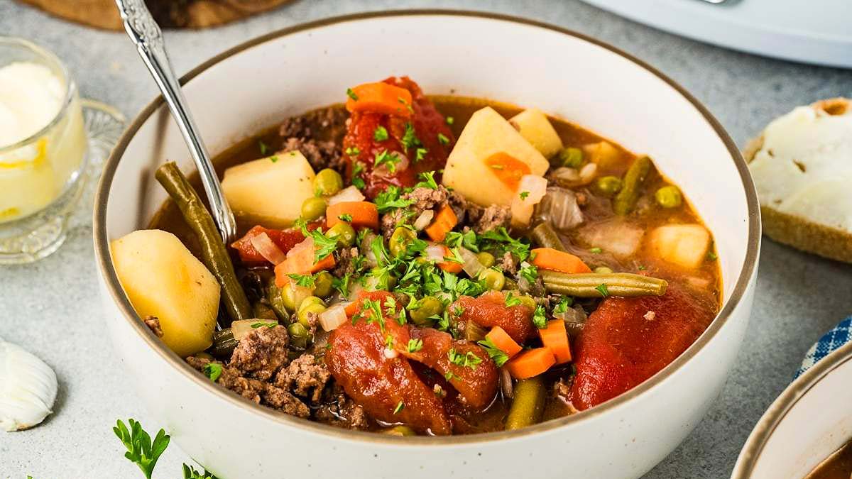 A bowl of beef and vegetable soup with potatoes, carrots, green beans, peas, tomatoes, and herbs, served with a spoon.