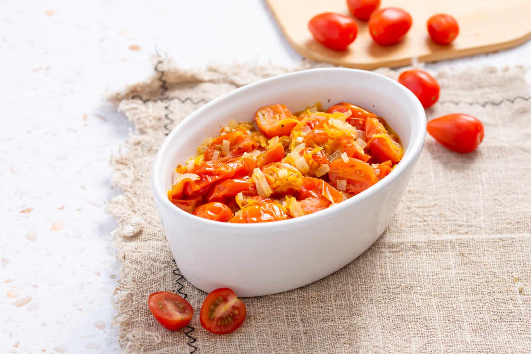 A white oval dish filled with a cooked rice and tomato mixture sits on a beige cloth, with fresh grape tomatoes scattered nearby.