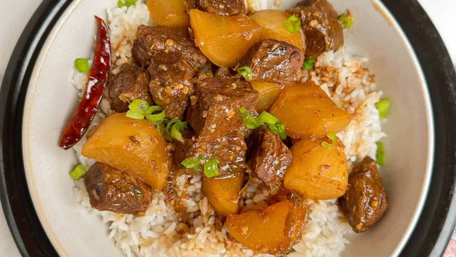 A bowl of white rice topped with chunks of braised beef, yellow potatoes, chopped green onions, and a whole dried red chili.