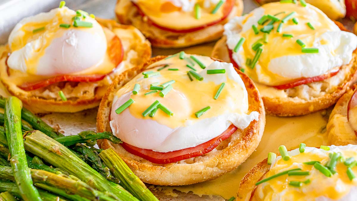 English muffins topped with ham, poached eggs, and hollandaise sauce, garnished with chopped chives, served with a side of roasted asparagus.