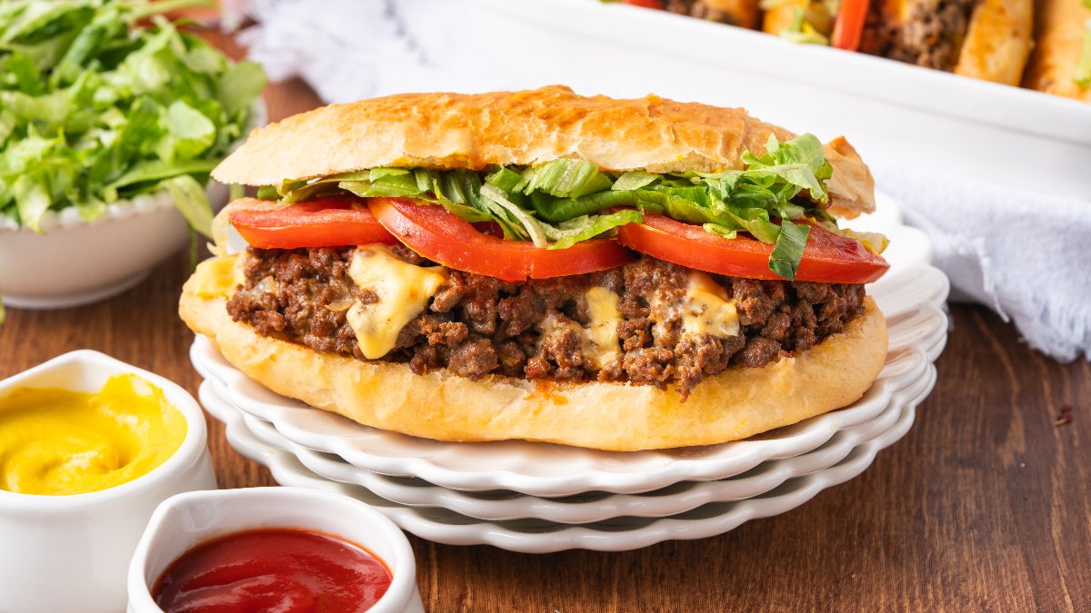 A sandwich with ground beef, melted cheese, tomato slices, and shredded lettuce on a hoagie roll, served on a white plate with ketchup, mustard, and lettuce on the side.