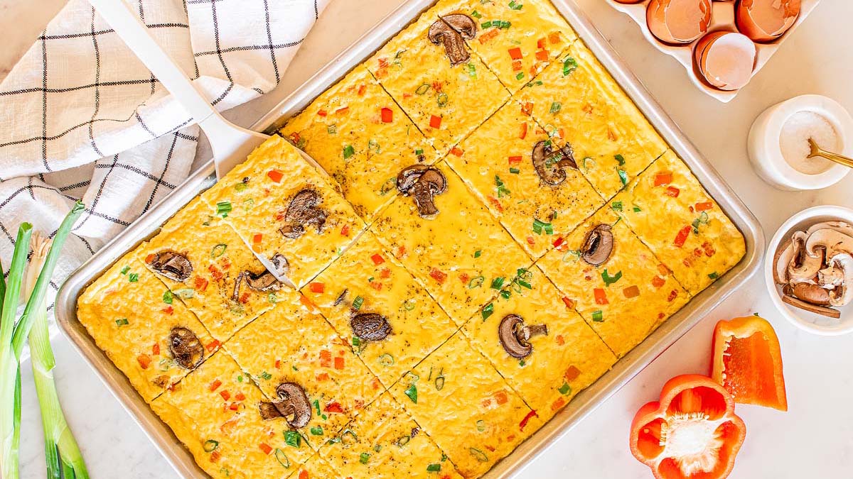 A baked sheet pan omelette with diced vegetables and mushrooms, sliced into squares, sits on a counter with eggs, scallions, mushrooms, and a red bell pepper nearby.