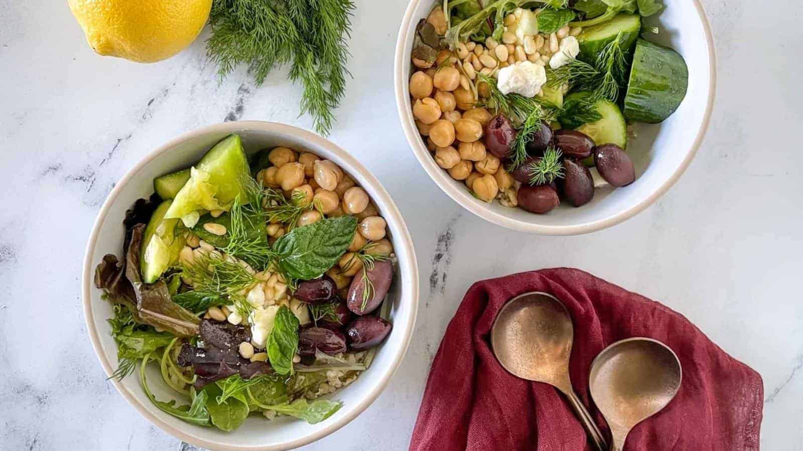 Two bowls of salad with chickpeas, olives, cucumbers, greens, and herbs on a white surface, next to a red napkin with two spoons and a lemon.