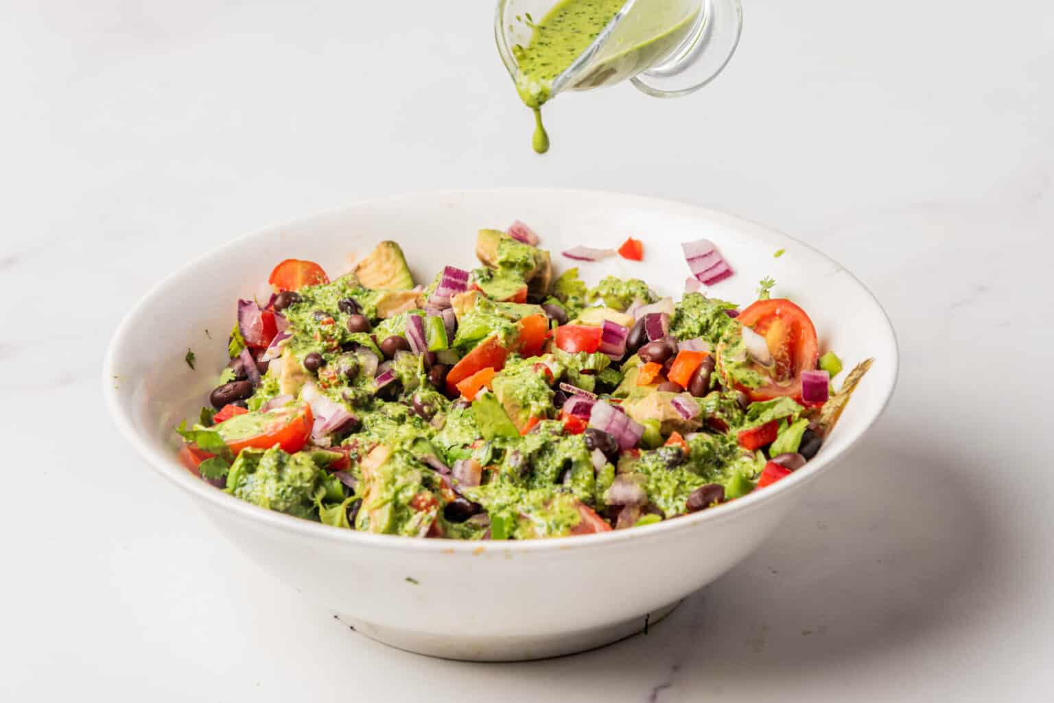 A white bowl filled with salad of tomatoes, beans, onions, and greens is being drizzled with green dressing from a small glass container.