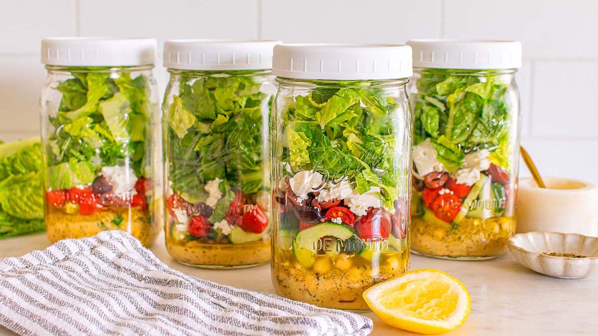 Four glass jars filled with layered salad ingredients, including lettuce, tomatoes, chickpeas, olives, and cheese, sit on a counter with salad dressing and a lemon nearby.