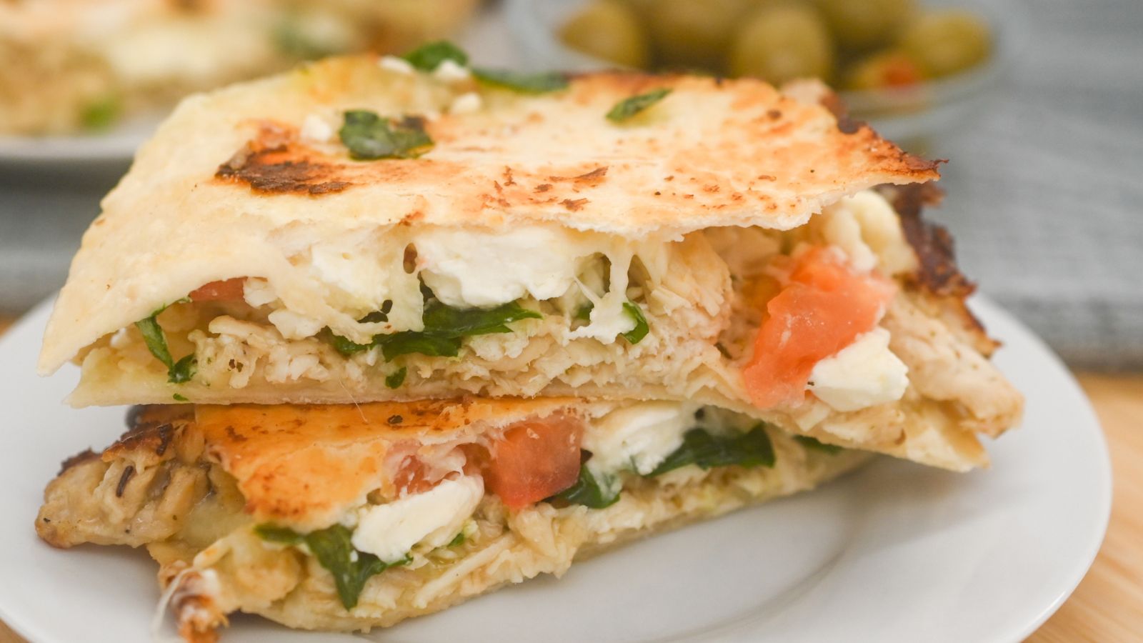 Two stacked slices of quesadilla filled with chicken, spinach, tomato, and cheese on a white plate, with a blurred bowl of olives in the background.