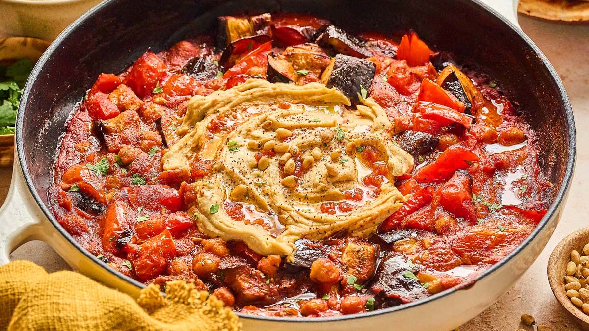 A skillet filled with a tomato-based stew containing chickpeas and vegetables, topped with a swirl of hummus and sprinkled with pine nuts.