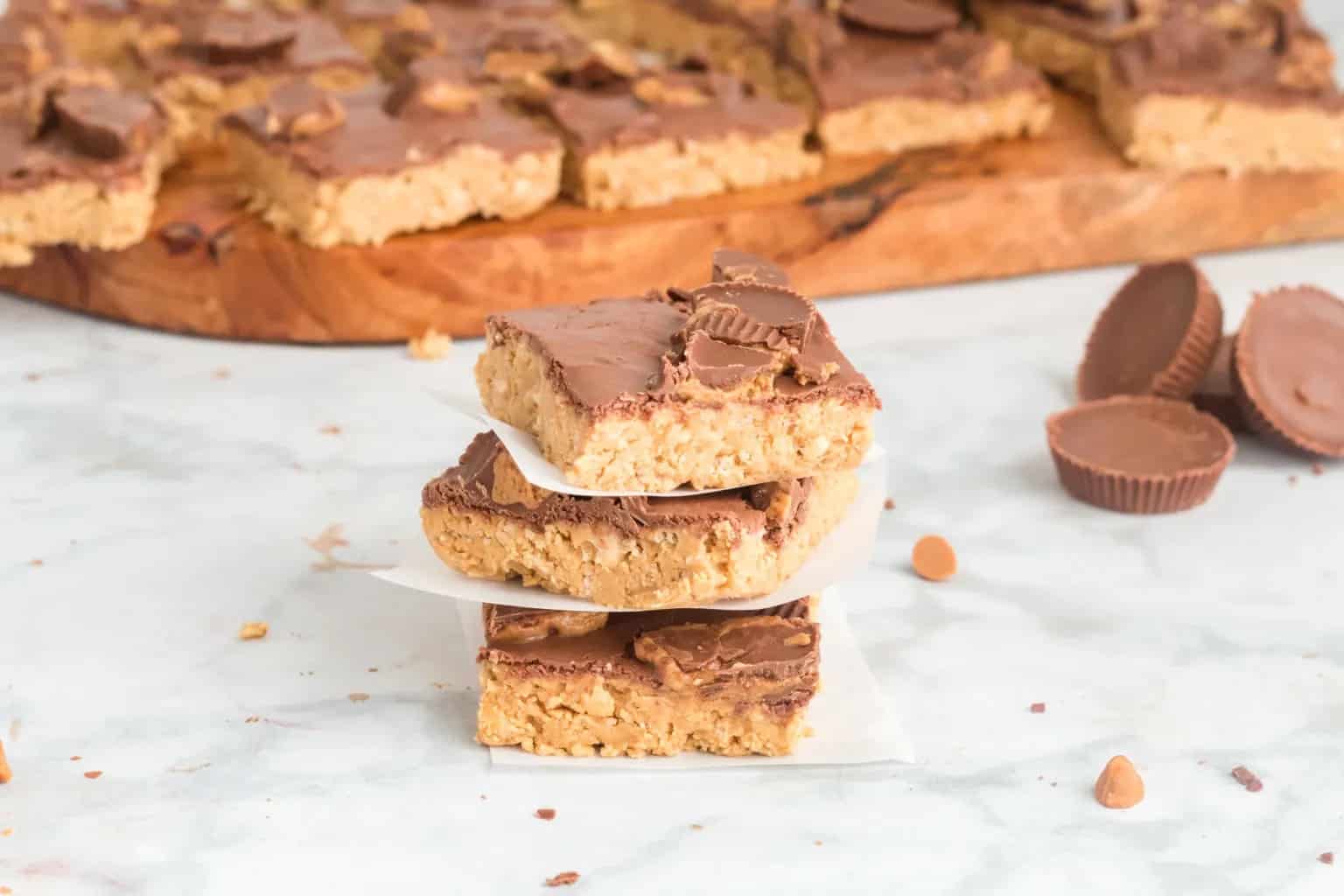 Three peanut butter chocolate bars are stacked in the foreground, with more bars and peanut butter cups visible on a wooden board in the background.