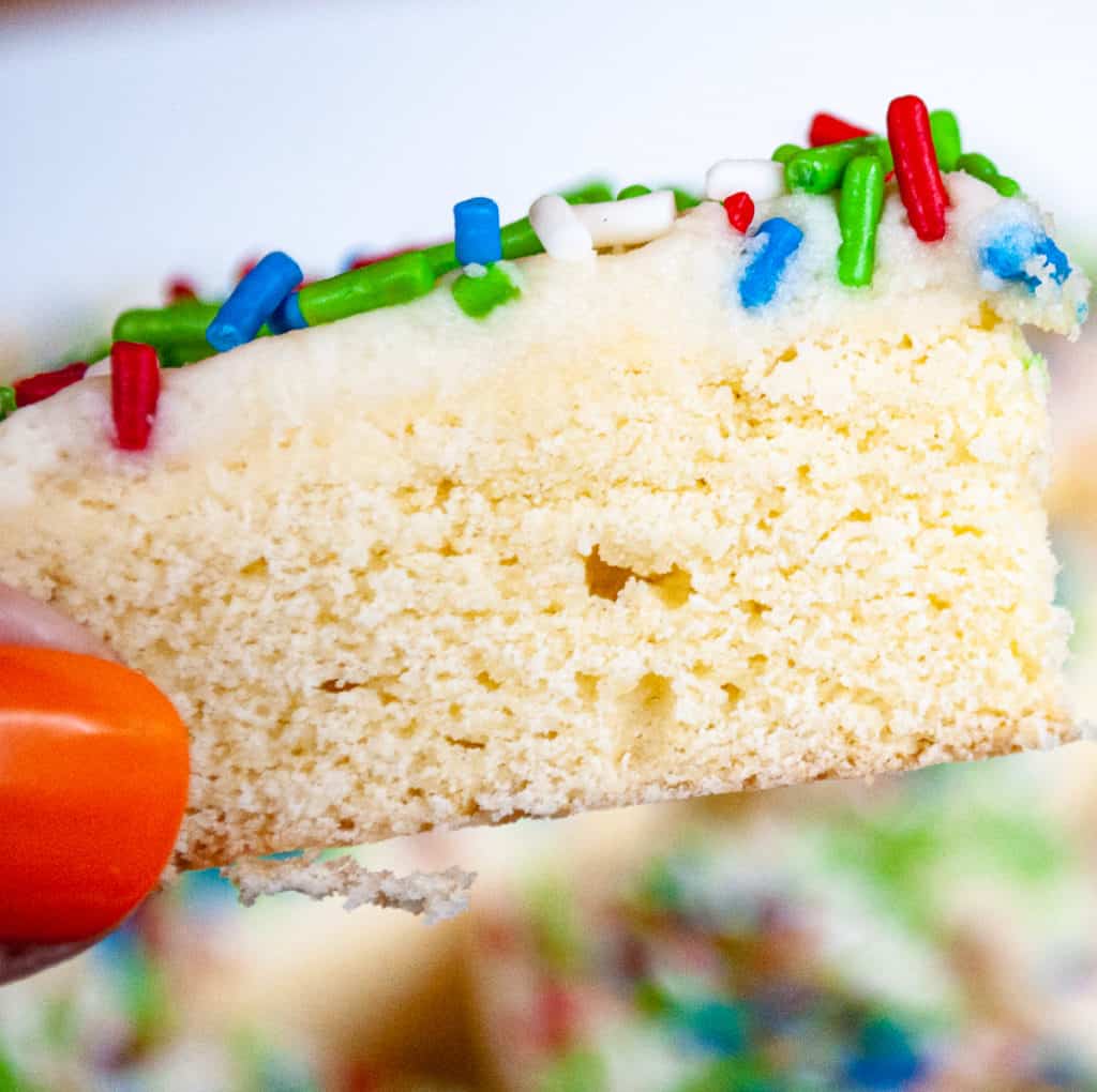 A close-up of a hand holding a thick slice of frosted sugar cookie topped with red, green, white, and blue sprinkles.