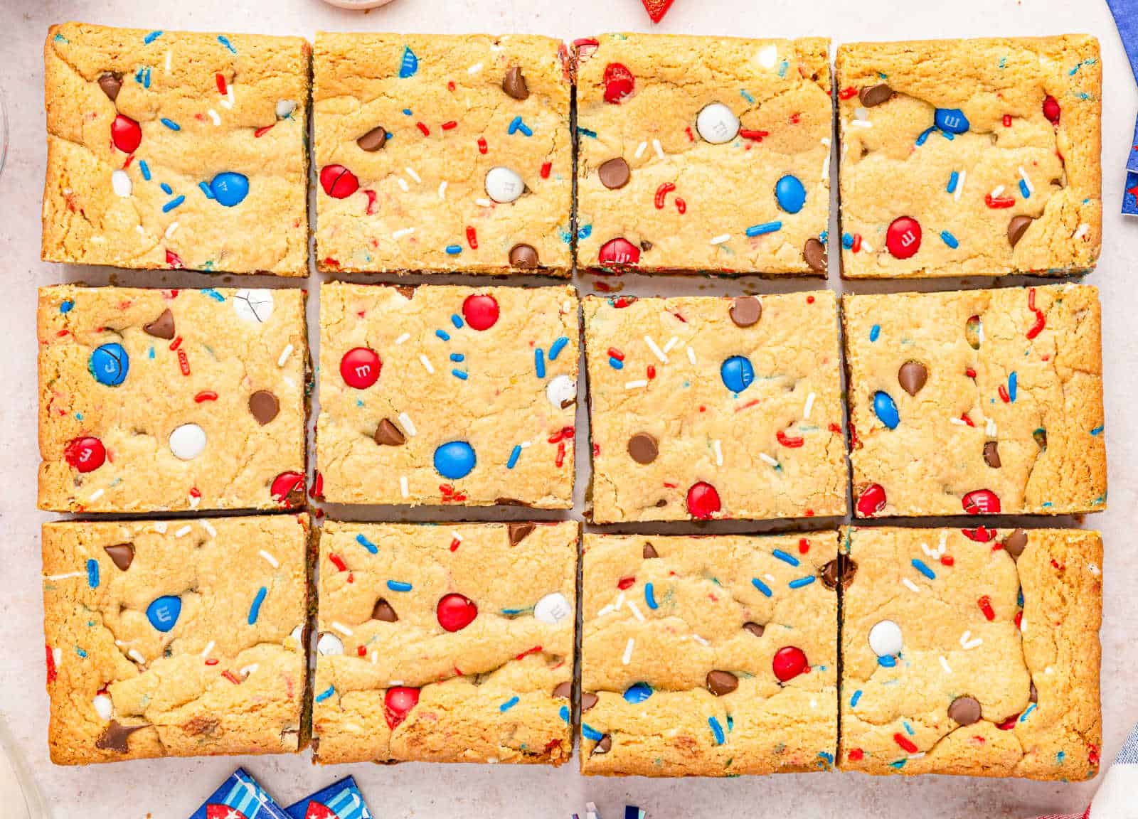 Twelve square cookie bars topped with red, white, and blue candy pieces, sprinkles, and chocolate chips arranged in a grid.