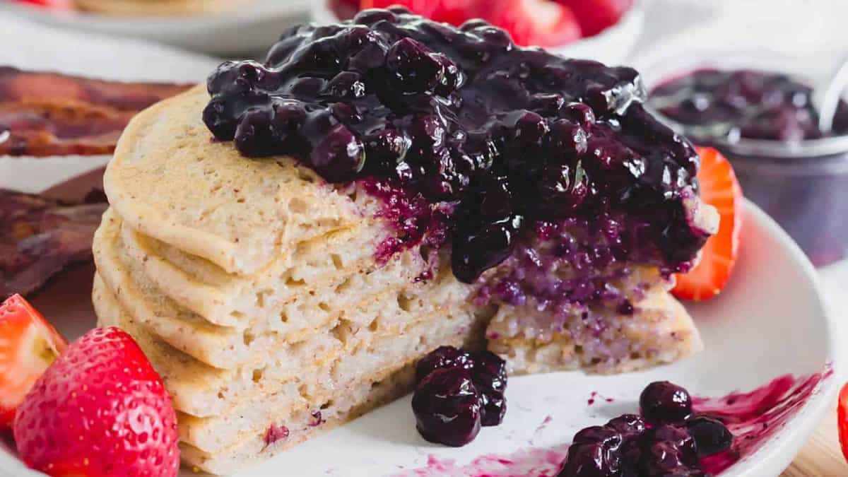 A stack of pancakes topped with blueberry compote, served with fresh strawberries and bacon on a white plate.