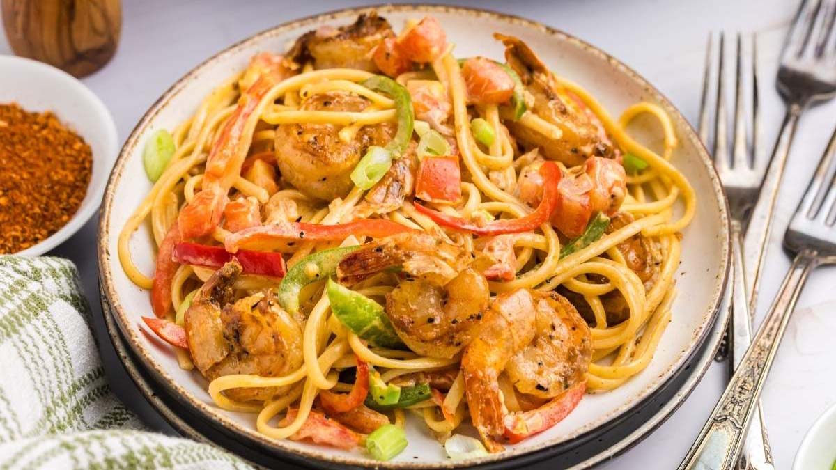 A plate of pasta with shrimp, sliced red bell peppers, green onions, and a creamy sauce, served with a fork and knife on the side.