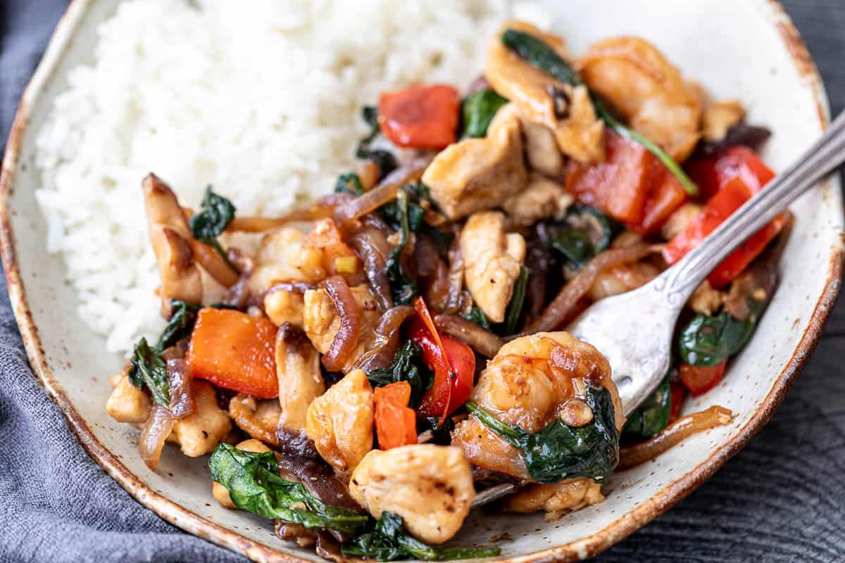 Plate of stir-fried chicken with shrimp, red bell peppers, onions, spinach, and white rice, with a fork resting on the dish.