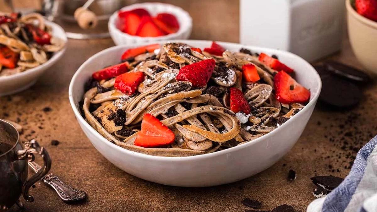 A bowl of pasta topped with sliced strawberries and crumbled cookies sits on a table, with more strawberries and cookies in bowls nearby.