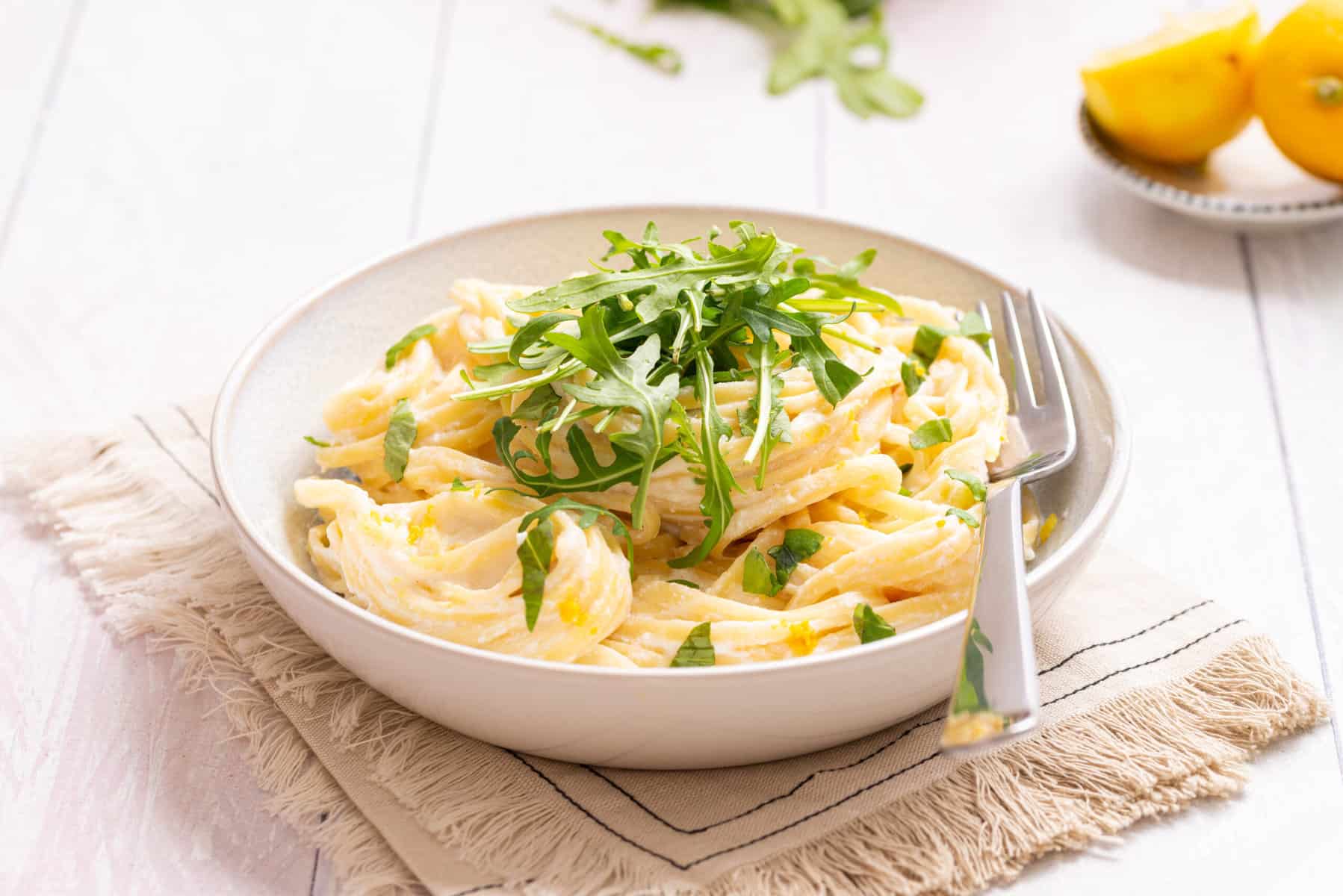 A bowl of creamy pasta garnished with fresh arugula is served with a fork on a neutral napkin, with lemon wedges and greens in the background.