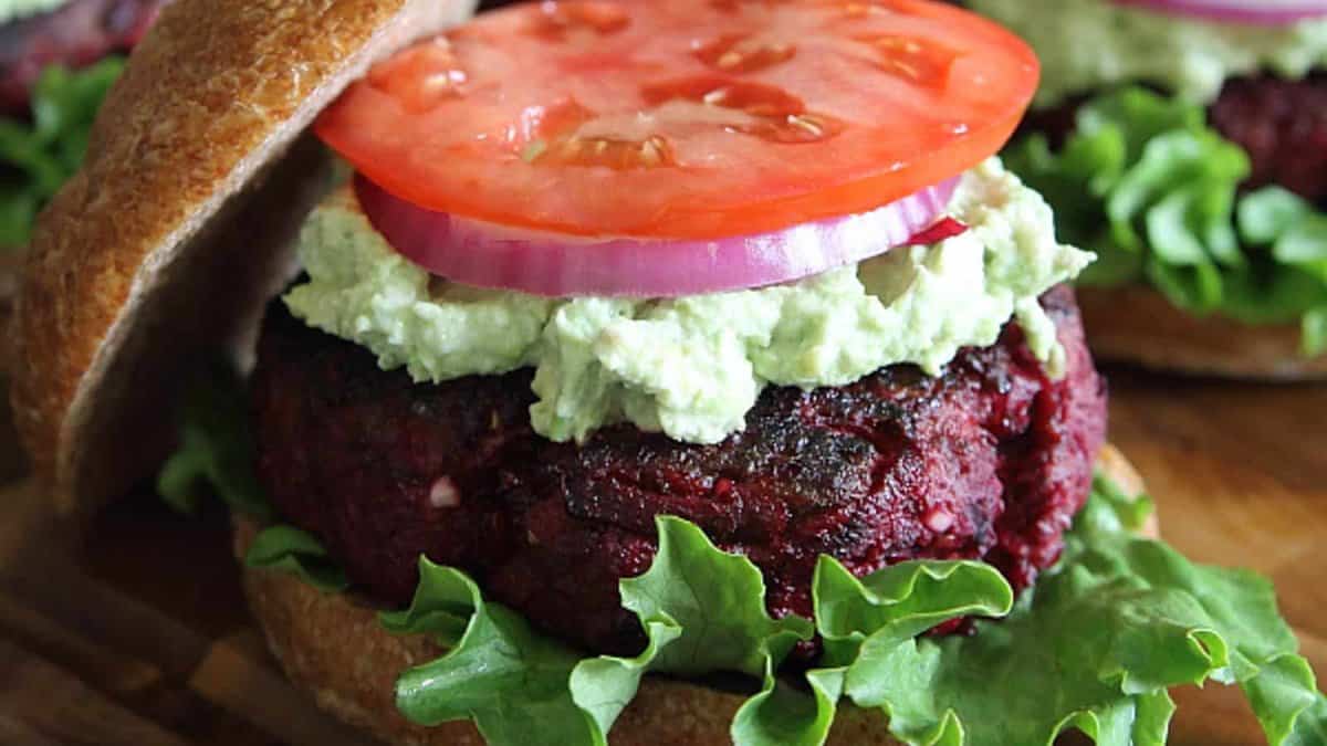 A veggie burger with lettuce, a beet-based patty, guacamole, sliced red onion, and a tomato slice on a whole wheat bun.