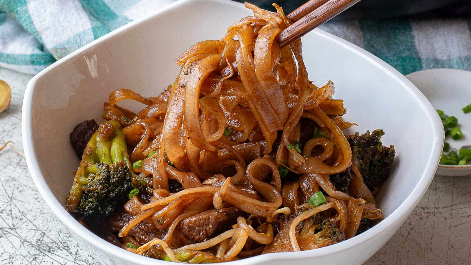 A bowl of stir-fried rice noodles with beef, broccoli, and bean sprouts, with chopsticks lifting a portion.
