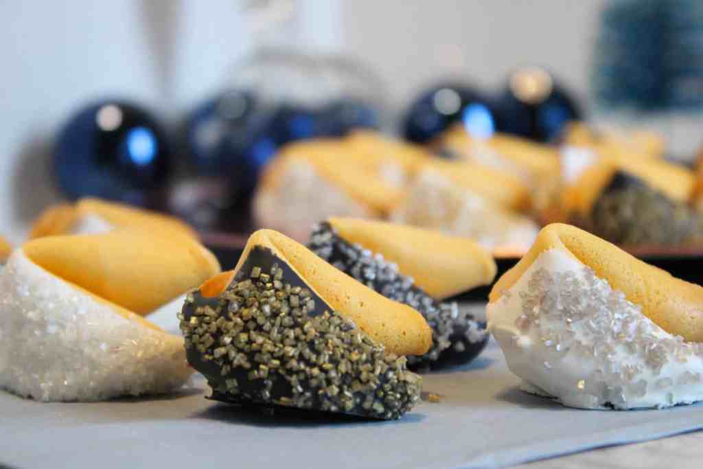 Several decorated fortune cookies are arranged on a table, each dipped in various coatings such as white, dark, and gold sprinkles.