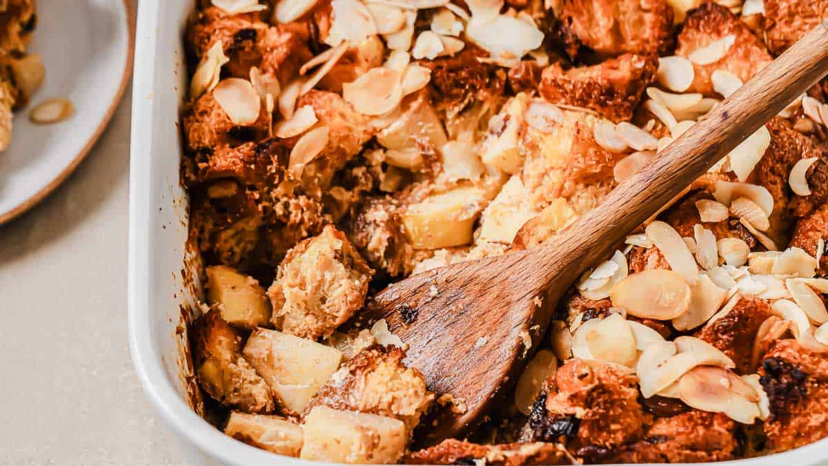 A baking dish filled with bread pudding mixed with apple chunks and topped with sliced almonds, with a wooden spoon resting inside.