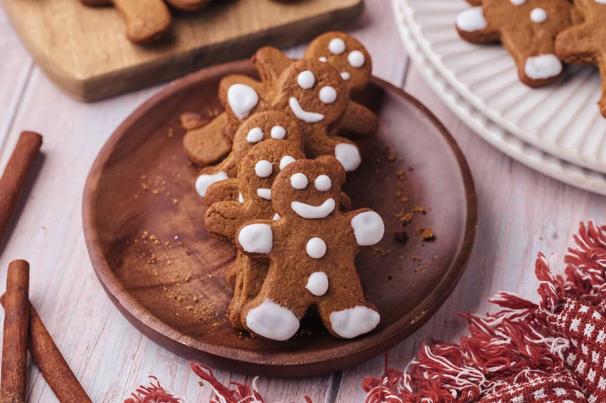 A wooden plate with several gingerbread cookies decorated with white icing sits on a light-colored table beside cinnamon sticks and a red fringed cloth.